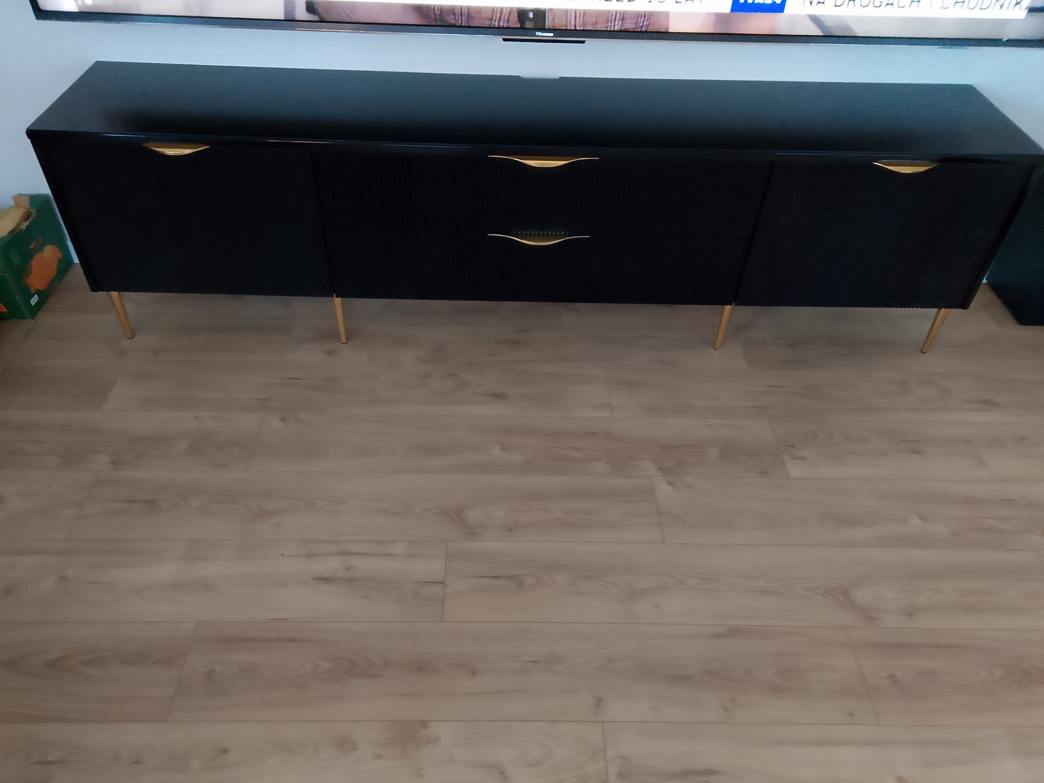 Czarna, lakierowana komoda z ryflowanymi frontami i złotymi uchwytami na cienkich, złotych nóżkach stoi na podłodze z paneli.