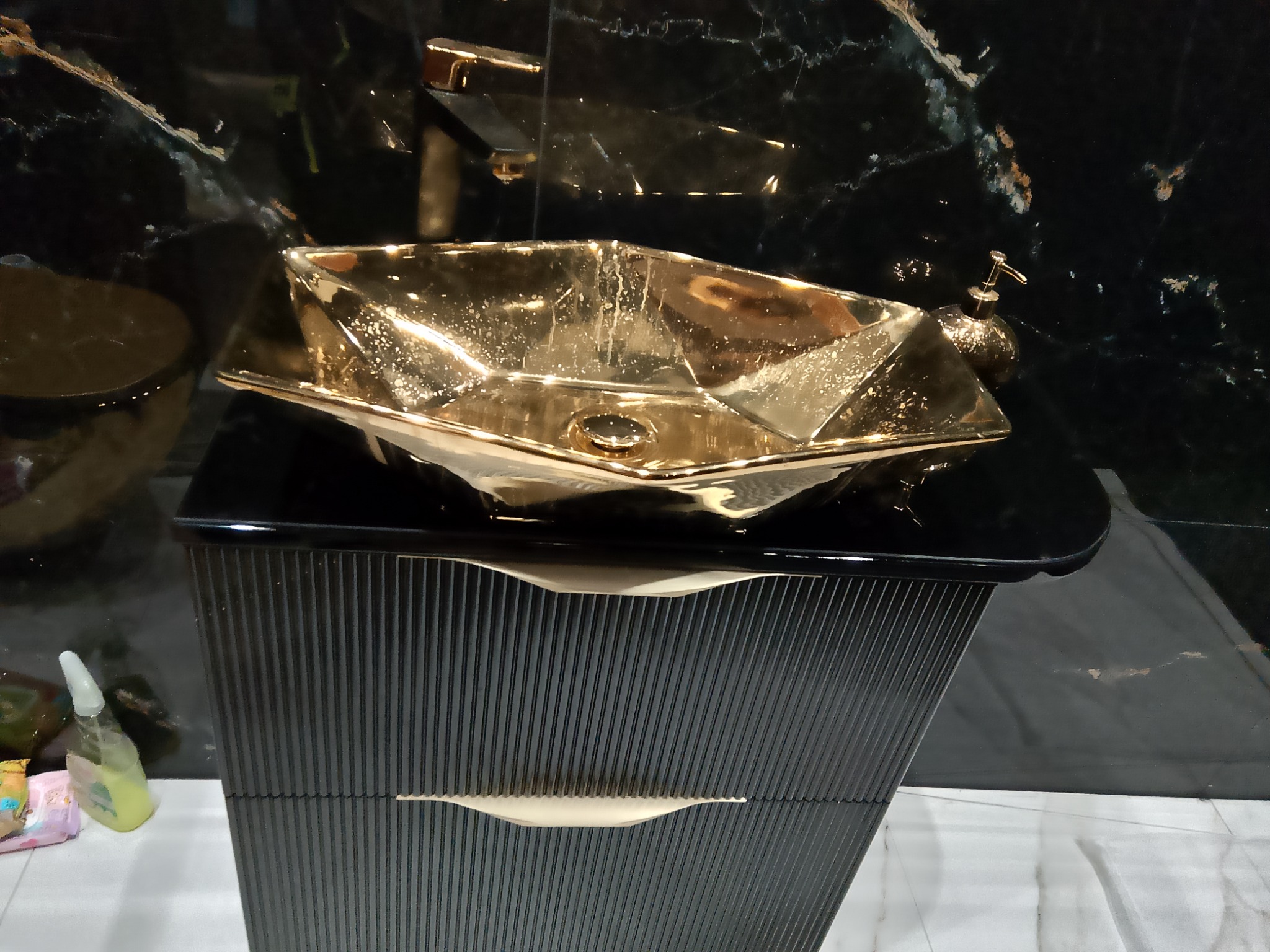 Złota, geometryczna umywalka nablatowa na czarnej szafce z ryflowanymi frontami i marmurowym tłem, z widoczną armaturą w kolorze złota.