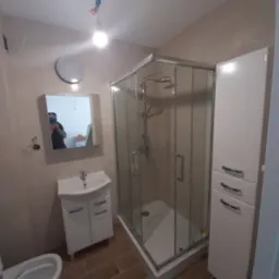 Wykończona łazienka z kabiną prysznicową narożną, umywalką z szafką, szafką stojącą i toaletą; brązowe płytki podłogowe imitujące drewno, beżowe płytki ścienne, w lustrze odbija się osoba w maseczce.