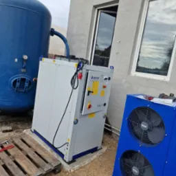 Zewnętrzna instalacja z niebieskim zbiornikiem ciśnieniowym i dwoma modułami chłodniczymi przy szarej ścianie budynku z oknami, widoczne narzędzia na palecie.