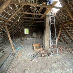 OliMax - Adaptacja strychu: mężczyzna na drabinie przy kominie, widoczne odsłonięte krokwie, stara cegła, deski podłogowe, okno w szczycie dachu i bałagan na podłodze.