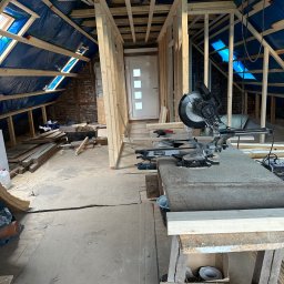 OliMax - Poddasze w trakcie adaptacji: widoczne drewniane konstrukcje ścian działowych i dachu pokrytego folią, okna dachowe wpuszczające światło, piła stołowa Scheppach na pierwszym planie, narzędzia...