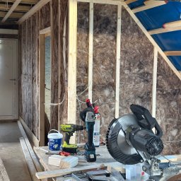 OliMax - Wnętrze w trakcie ocieplania wełną mineralną, widoczne narzędzia: ukośnica Scheppach, wiertarka akumulatorowa, pistolet do pianki montażowej oraz wiadro z klejem do płyt gipsowych, częściowo...