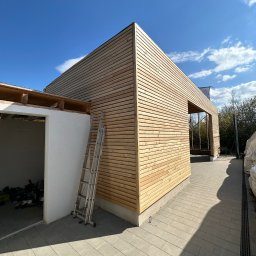 OliMax - Budynek w trakcie wykańczania elewacji drewnianą boazerią, z widoczną drabiną i fragmentem surowego wnętrza, na tle błękitnego nieba.