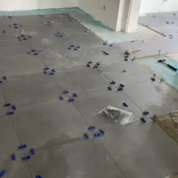 Szare płytki podłogowe w trakcie układania, z użyciem niebieskich klipsów poziomujących, widoczny klej, paczka klipsów, młotek gumowy i wiadro z zaprawą.