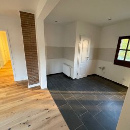 OliMax - Wnętrze połączone z dwoma rodzajami podłóg: drewnianą i z szarych płytek. Widoczny fragment ściany z cegły, białe ściany z szarym paskiem, okno i grzejnik.