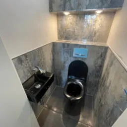 Nowoczesna, wykończona łazienka w Rumii z czarną ceramiką: toaleta i umywalka, szare płytki imitujące kamień na ścianach i podłodze, oświetlenie punktowe.