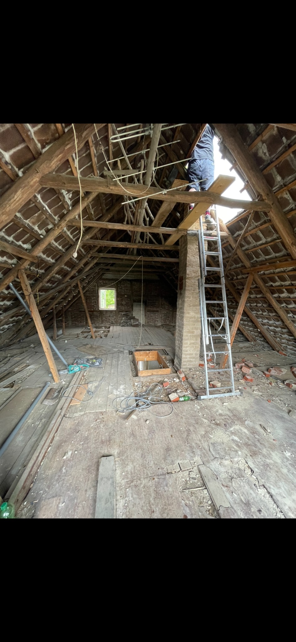 Adaptacja strychu: mężczyzna na drabinie przy kominie, widoczne odsłonięte krokwie, stara cegła, deski podłogowe, okno w szczycie dachu i bałagan na podłodze.