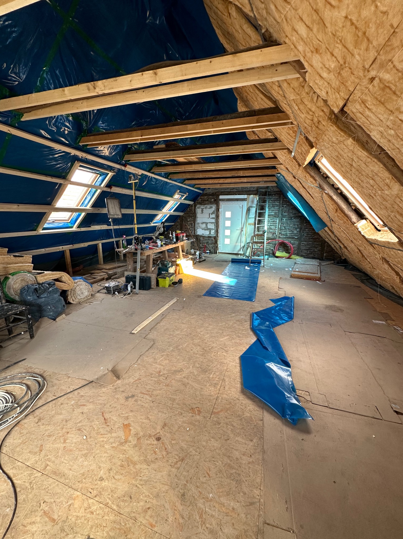 Poddasze w trakcie adaptacji w Rumi, widoczne ocieplenie ścian wełną mineralną, folia paroizolacyjna na krokwiach, okna dachowe, narzędzia i materiały budowlane przygotowane do dalszych prac.