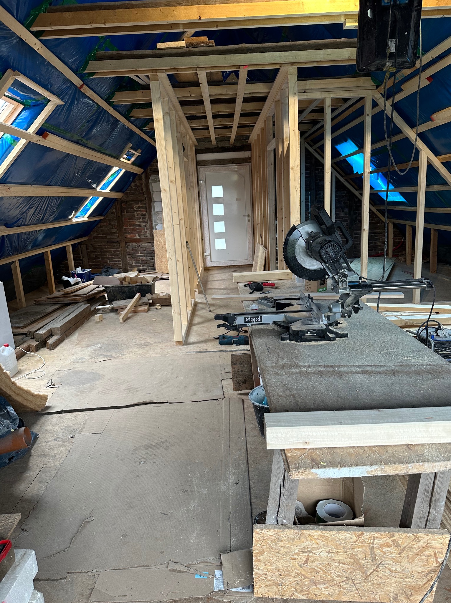 Poddasze w trakcie adaptacji: widoczne drewniane konstrukcje ścian działowych i dachu pokrytego folią, okna dachowe wpuszczające światło, piła stołowa Scheppach na pierwszym planie, narzędzia...