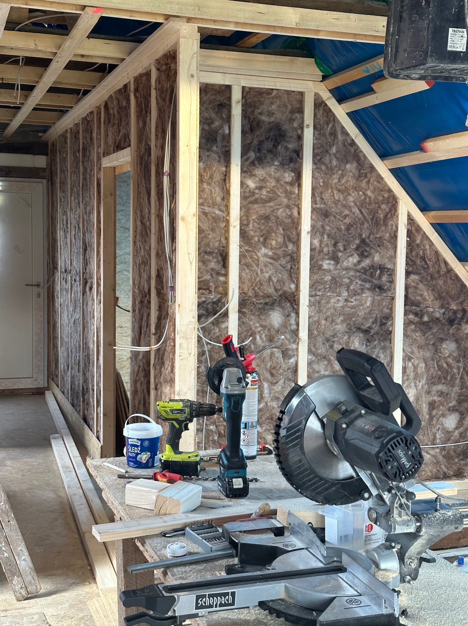 Wnętrze w trakcie ocieplania wełną mineralną, widoczne narzędzia: ukośnica Scheppach, wiertarka akumulatorowa, pistolet do pianki montażowej oraz wiadro z klejem do płyt gipsowych, częściowo...