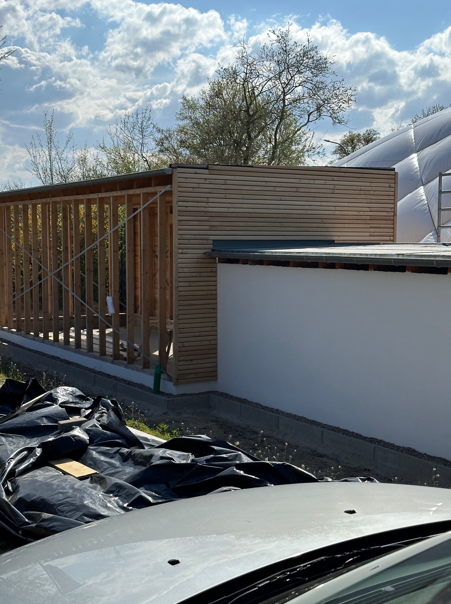 Konstrukcja drewniana w trakcie budowy, częściowo obłożona panelami drewnianymi, widoczne czarne folie budowlane i fragment auta w tle.