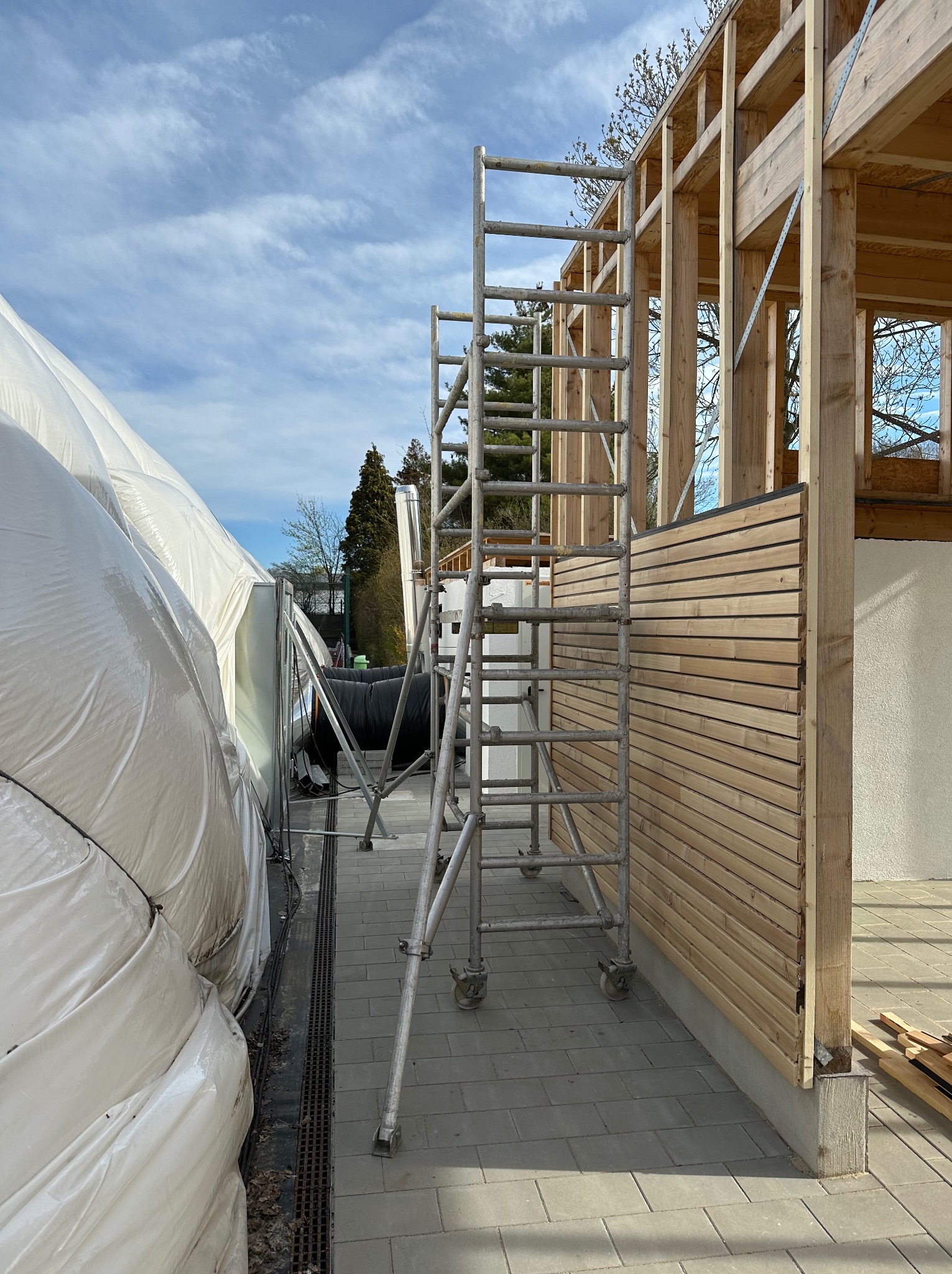 Wznoszenie drewnianej konstrukcji budynku z zamontowaną częścią elewacji z desek, widoczne rusztowanie i zabezpieczająca folia.