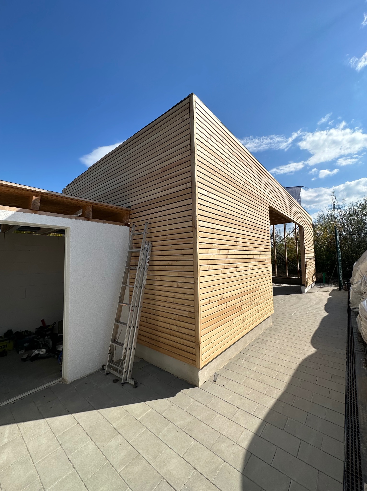 Budynek w trakcie wykańczania elewacji drewnianą boazerią, z widoczną drabiną i fragmentem surowego wnętrza, na tle błękitnego nieba.