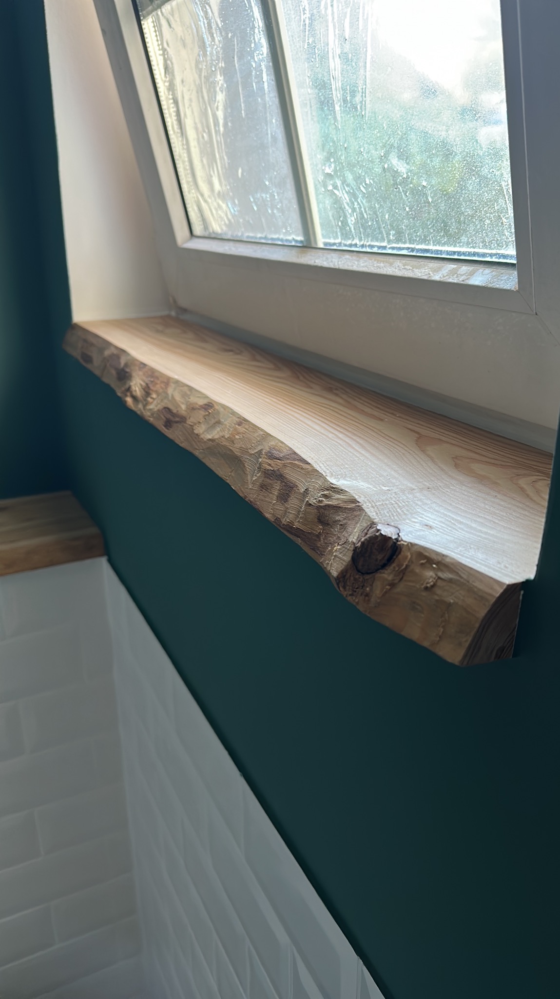 Półka z surowego drewna z naturalną krawędzią zamontowana pod oknem na poddaszu w łazience z białą, kafelkową ścianą i ciemnozieloną ścianą boczną.