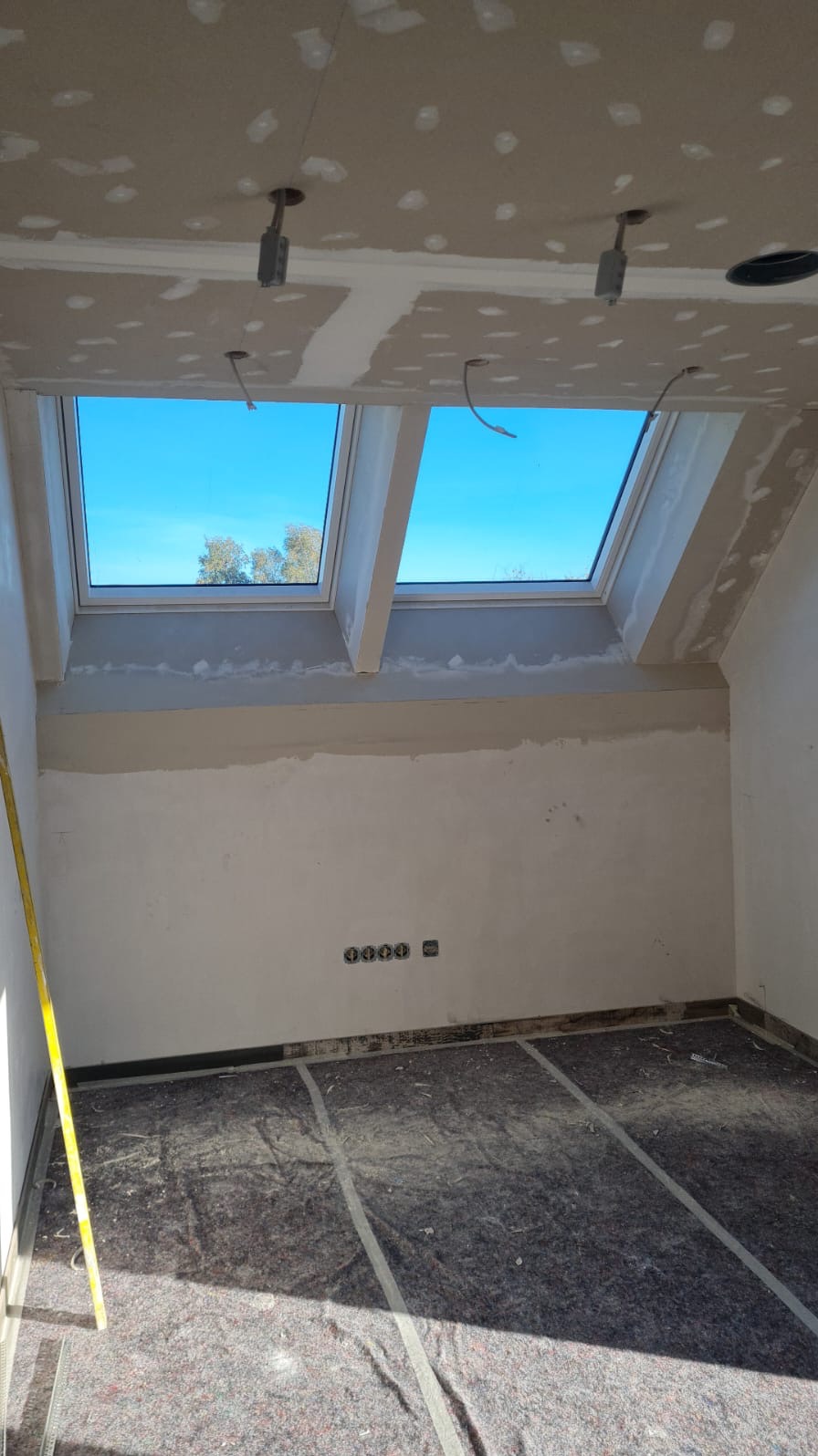 Pokój w trakcie prac wykończeniowych: widoczne dwa okna dachowe, ściany po szpachlowaniu, zabezpieczona podłoga, wystające przewody elektryczne i poziomica oparta o ścianę.