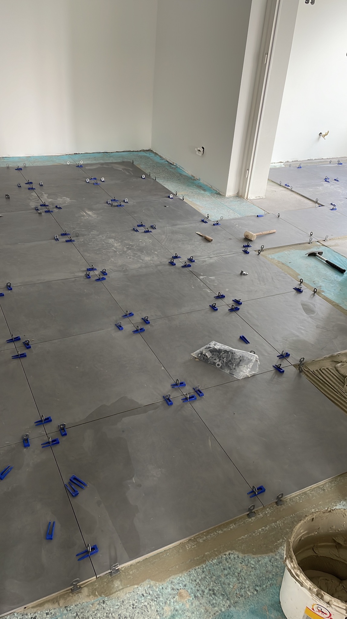 Szare płytki podłogowe w trakcie układania, z użyciem niebieskich klipsów poziomujących, widoczny klej, paczka klipsów, młotek gumowy i wiadro z zaprawą.