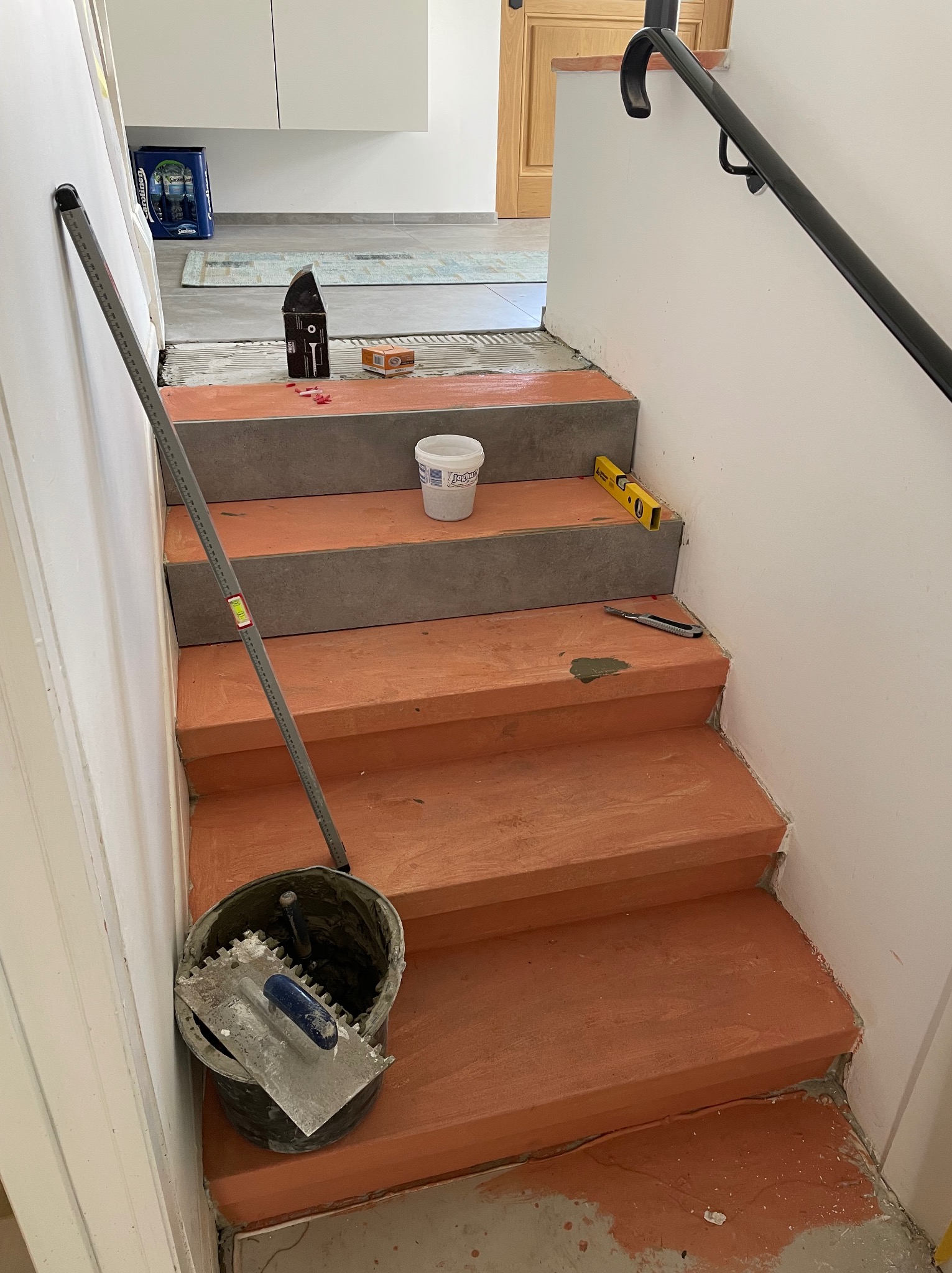 Układanie płytek na schodach wewnętrznych: widoczny etap prac z użyciem poziomicy, wiadra z narzędziami (szpachelka ząbkowana), listwy pomiarowej i nożyka do cięcia płytek. Część schodów wykończona...