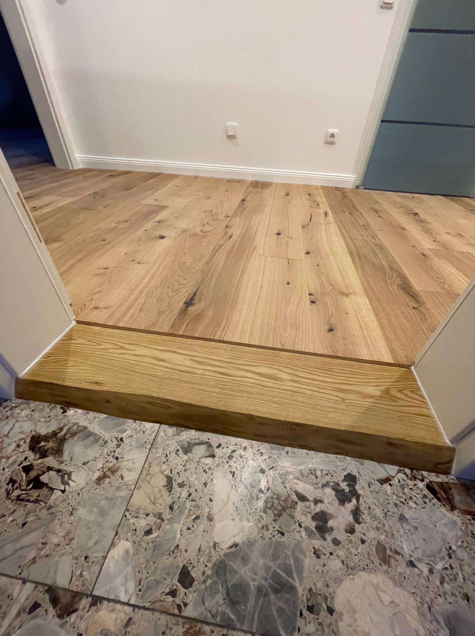 Jasne panele podłogowe z drewna, przechodzące przez drewniany próg do marmurowej posadzki, widok z góry na łączenie dwóch rodzajów podłóg.