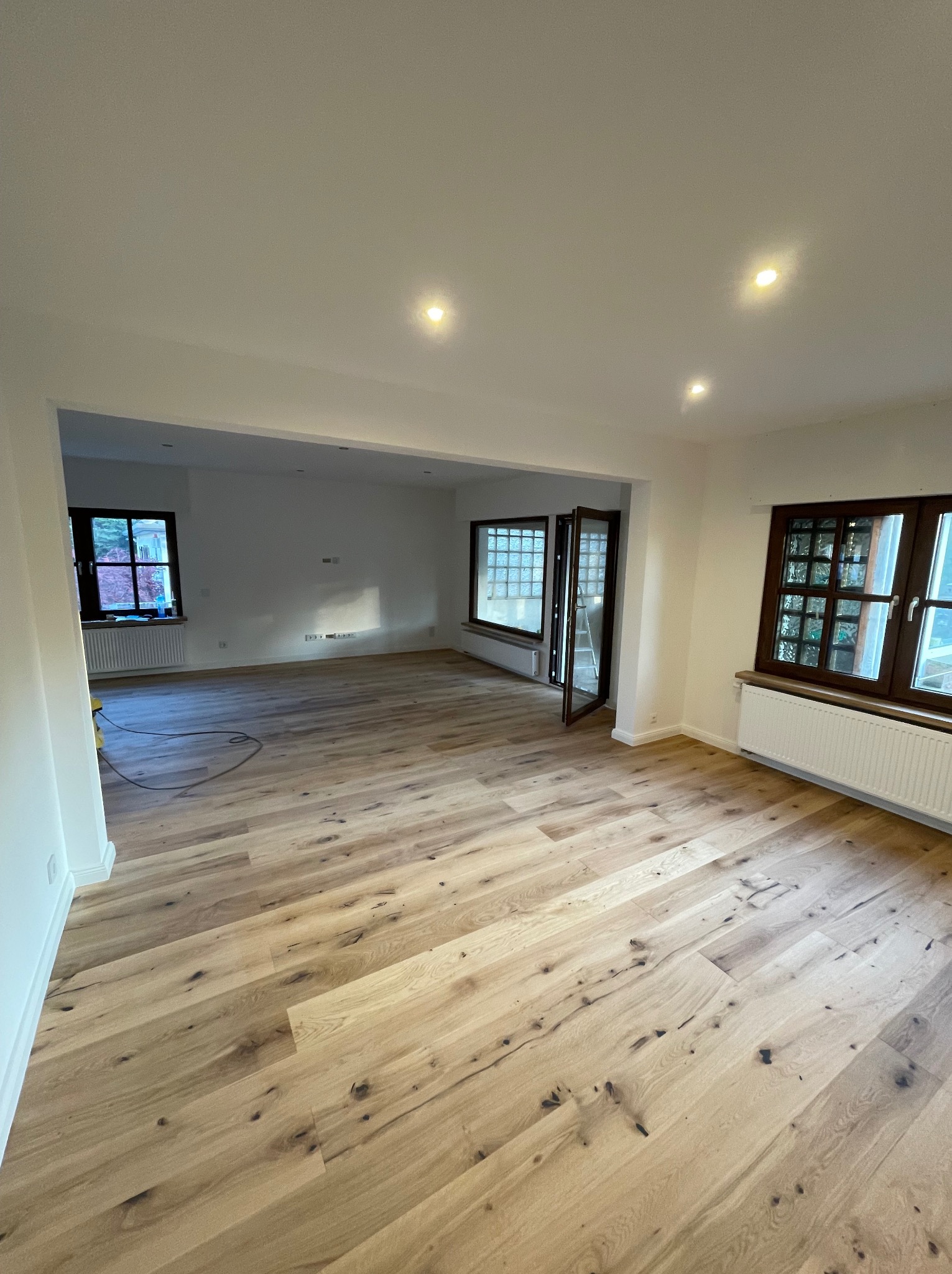 Jasne panele podłogowe ułożone w przestronnym, odnowionym salonie z białymi ścianami i oknami z ciemnymi ramami. Widoczny fragment żółtego odkurzacza w rogu pomieszczenia.