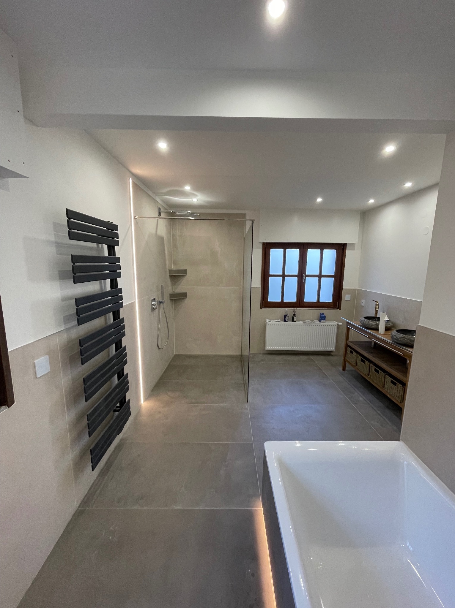 Nowoczesna łazienka z szarą podłogą, przeszkloną kabiną prysznicową, grzejnikiem drabinkowym w kolorze antracytowym, drewnianą szafką z umywalkami i oknem z drewnianą ramą.