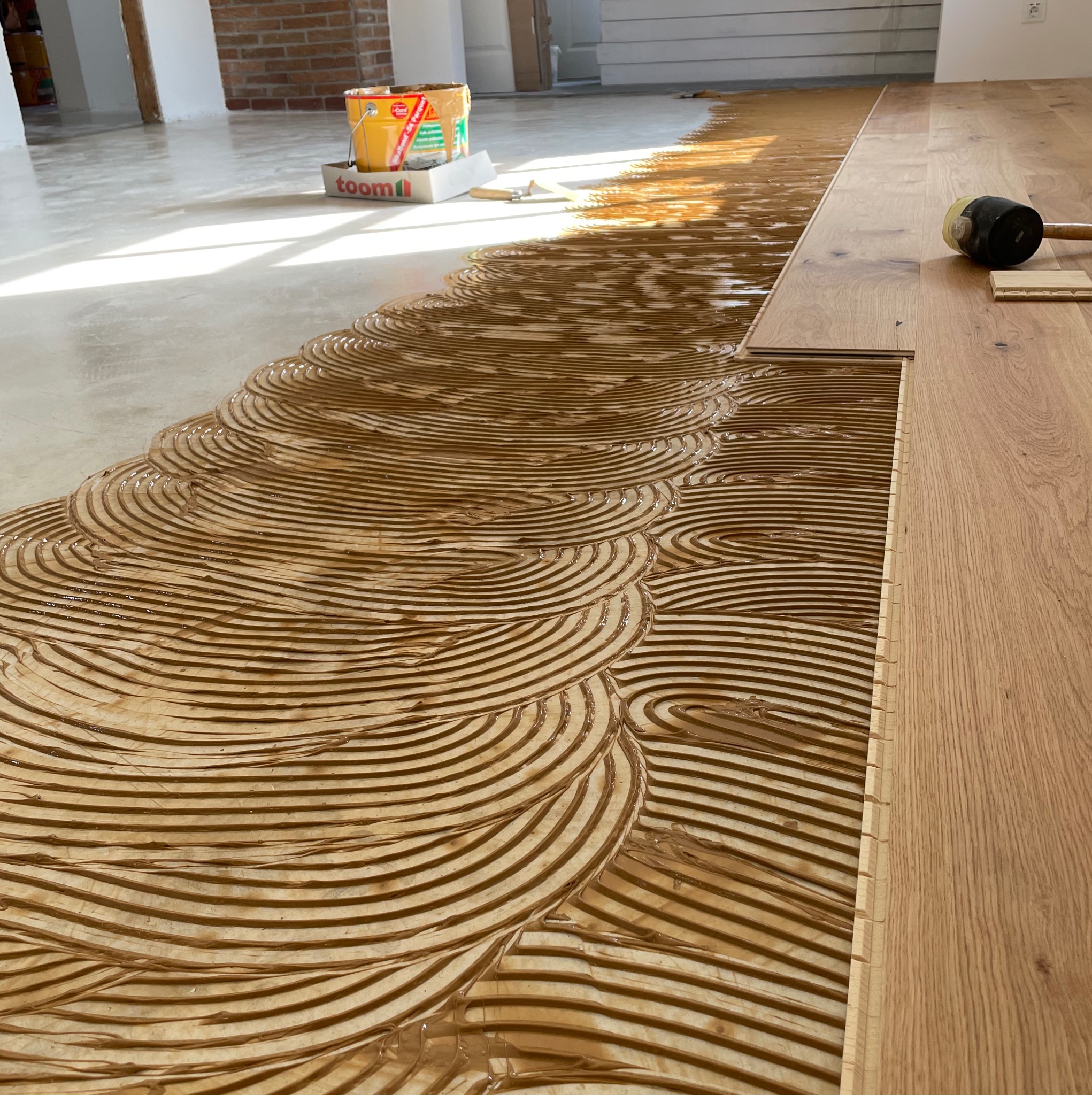 Układanie paneli podłogowych: świeżo nałożony klej w charakterystyczne półkola, widoczna część ułożonej podłogi i gumowy młotek.