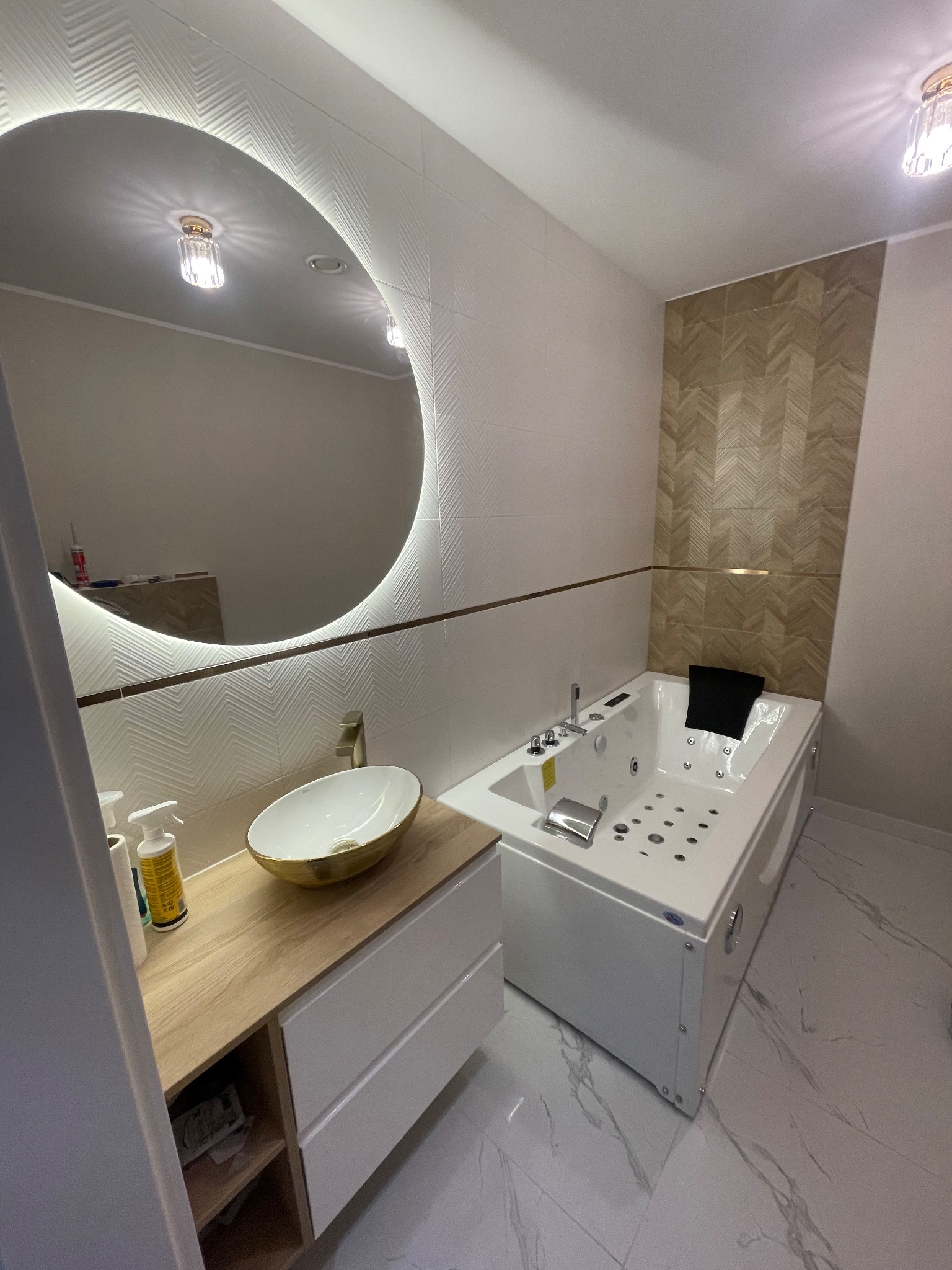 Nowoczesna łazienka z owalną umywalką w kolorze białym ze złotym obramowaniem, podświetlanym okrągłym lustrem, białą wanną z hydromasażem i dekoracyjną ścianą z płytek w odcieniach beżu.