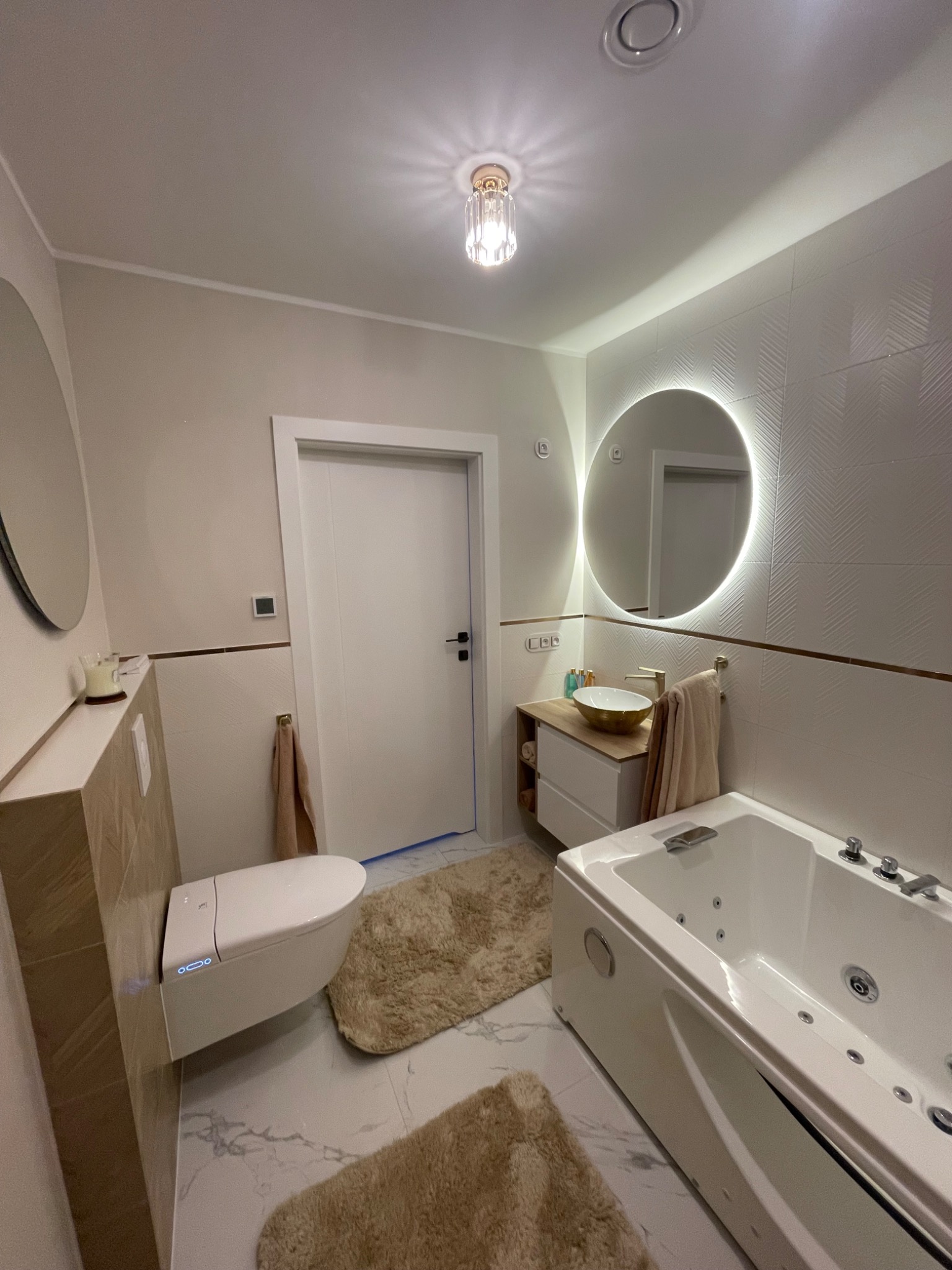 Nowoczesna łazienka z wiszącą toaletą z podświetleniem LED, okrągłym lustrem z podświetleniem, białą wanną z hydromasażem i beżowymi dywanikami.