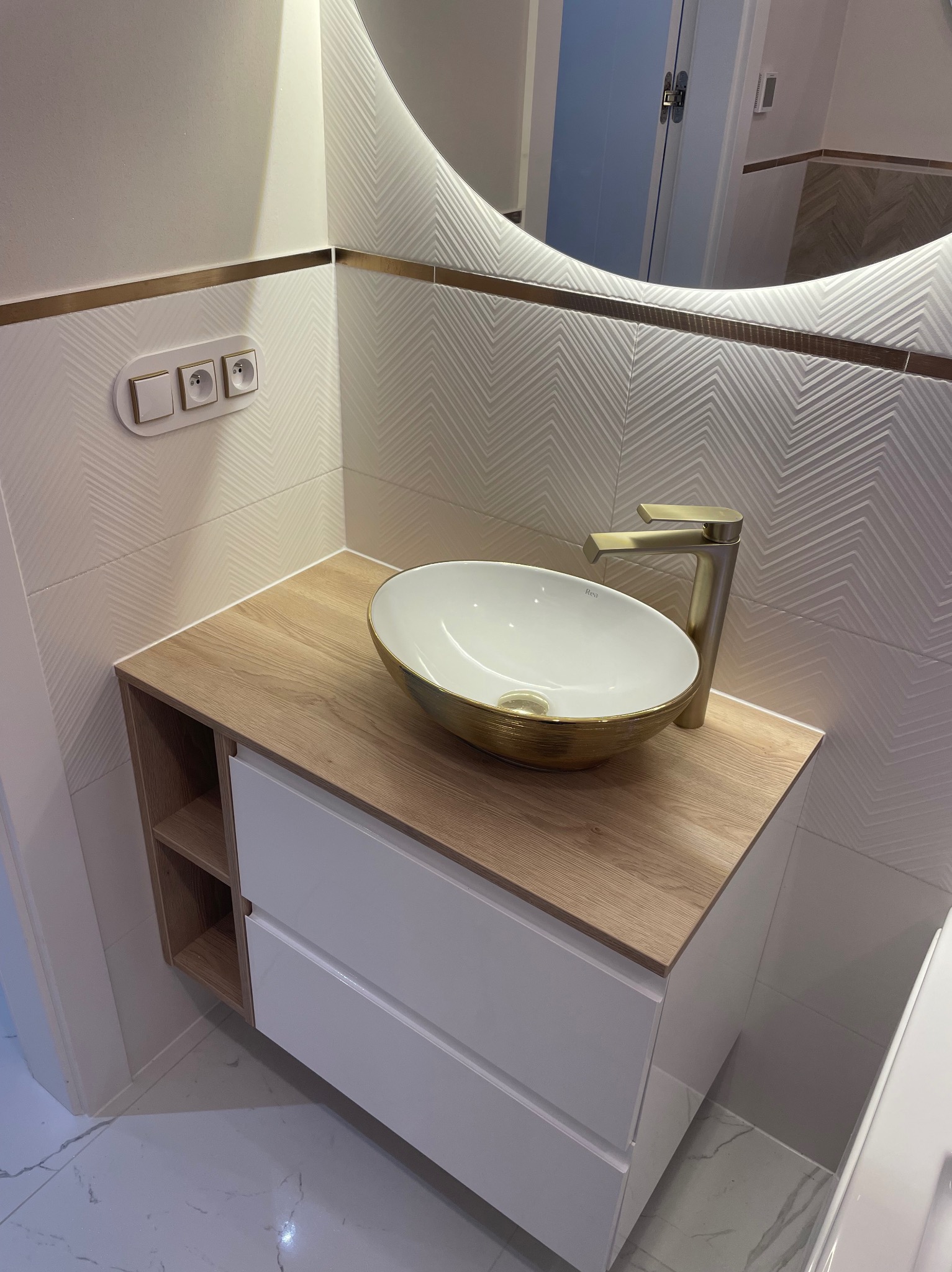 Nowoczesna łazienka z owalną umywalką w kolorze białym ze złotym obramowaniem, złotą baterią, szafką z drewnianym blatem i białymi frontami, oraz płytkami w geometryczny wzór z delikatnym złotym...