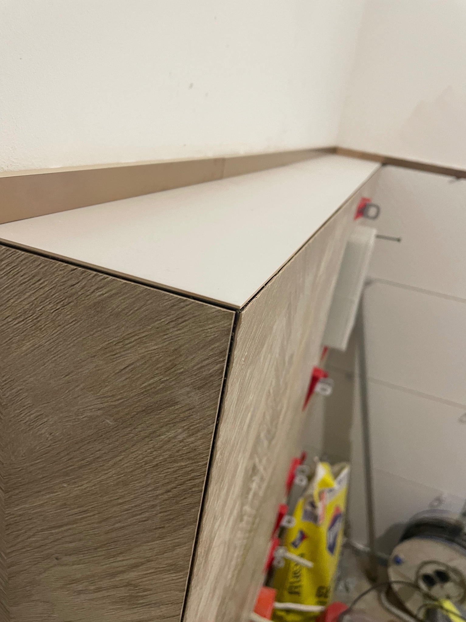 Narożny fragment ściany wyłożonej białymi płytkami ceramicznymi z widocznymi klipsami poziomującymi, sąsiadujący z zabudową w kolorze jasnego drewna, z elementami wyposażenia w tle.