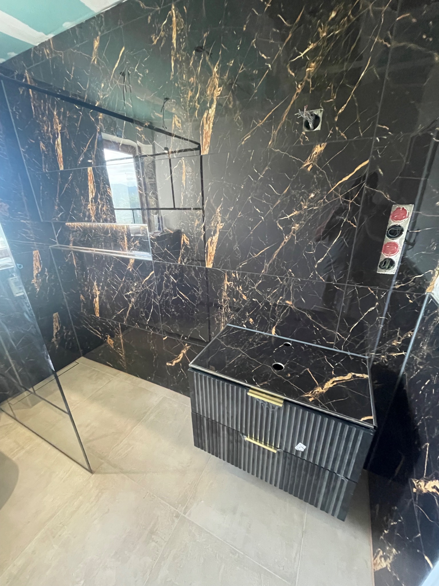 Nowoczesna łazienka z czarnymi płytkami imitującymi marmur ze złotymi żyłami, wisząca szafka pod umywalkę z ryflowanymi frontami i złotymi uchwytami, widoczne otwarte przewody elektryczne...