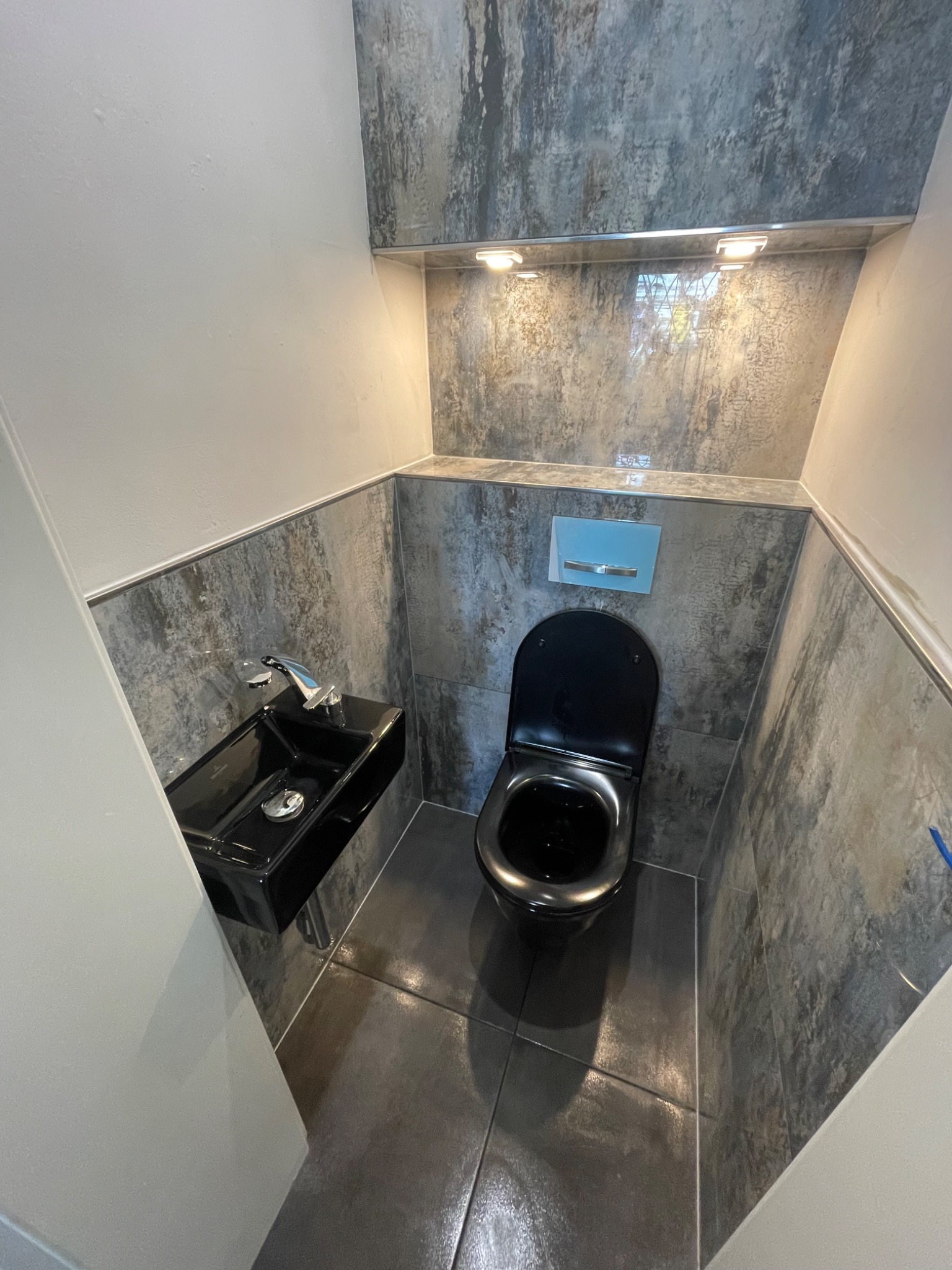 Nowoczesna, wykończona łazienka w Rumii z czarną ceramiką: toaleta i umywalka, szare płytki imitujące kamień na ścianach i podłodze, oświetlenie punktowe.