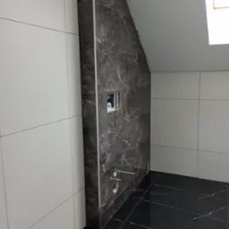 Wykończona łazienka na poddaszu z czarną marmurową podłogą i ścianą, białe płytki na ścianach bocznych, widoczne otwory na baterię i instalację sanitarną.