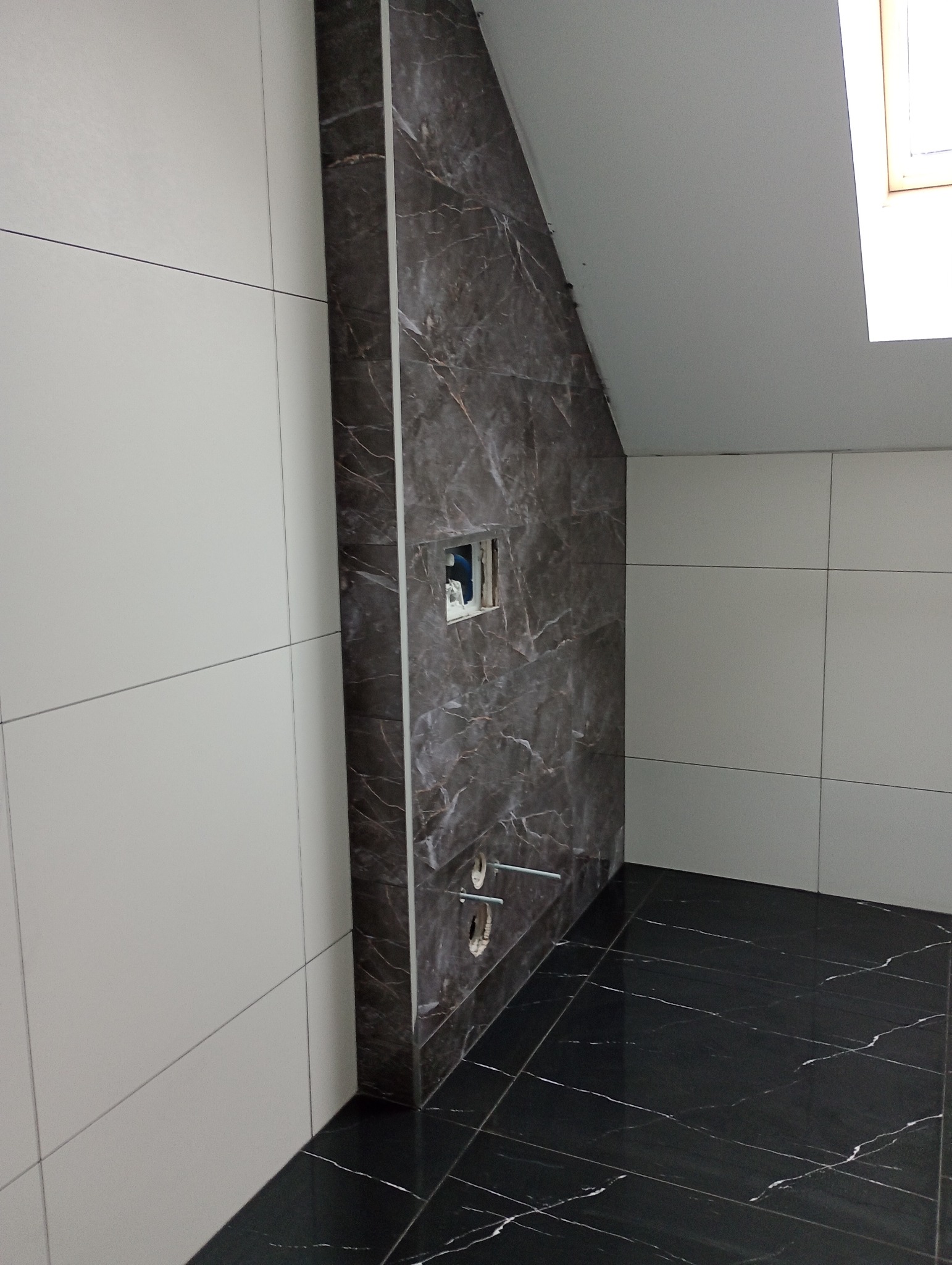 Wykończona łazienka na poddaszu z czarną marmurową podłogą i ścianą, białe płytki na ścianach bocznych, widoczne otwory na baterię i instalację sanitarną.