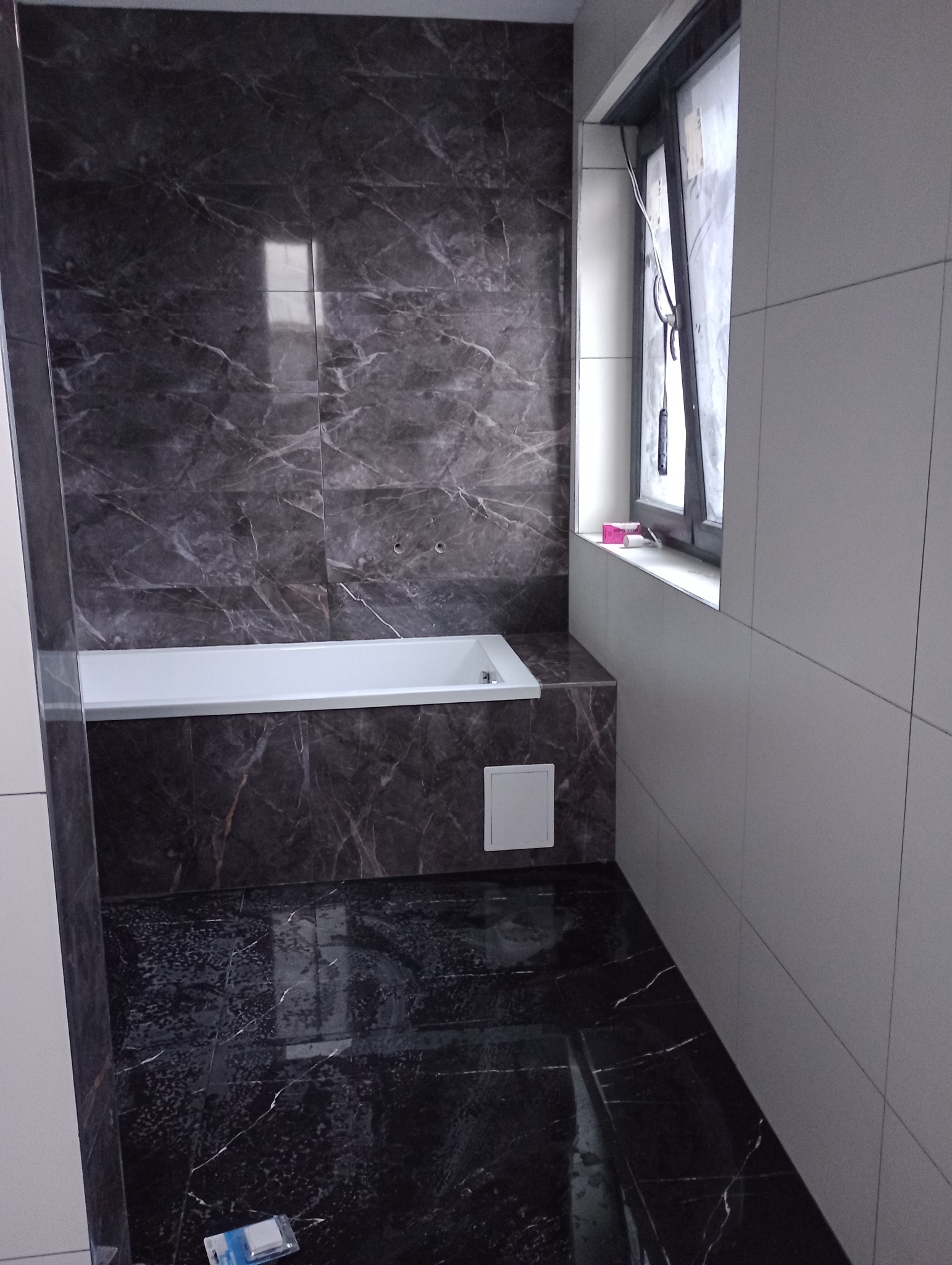 Łazienka z wanną obłożoną ciemnymi płytkami imitującymi marmur, widoczna biała wanna, okno i fragment podłogi z podobnych płytek, mała klapka rewizyjna w obudowie wanny.