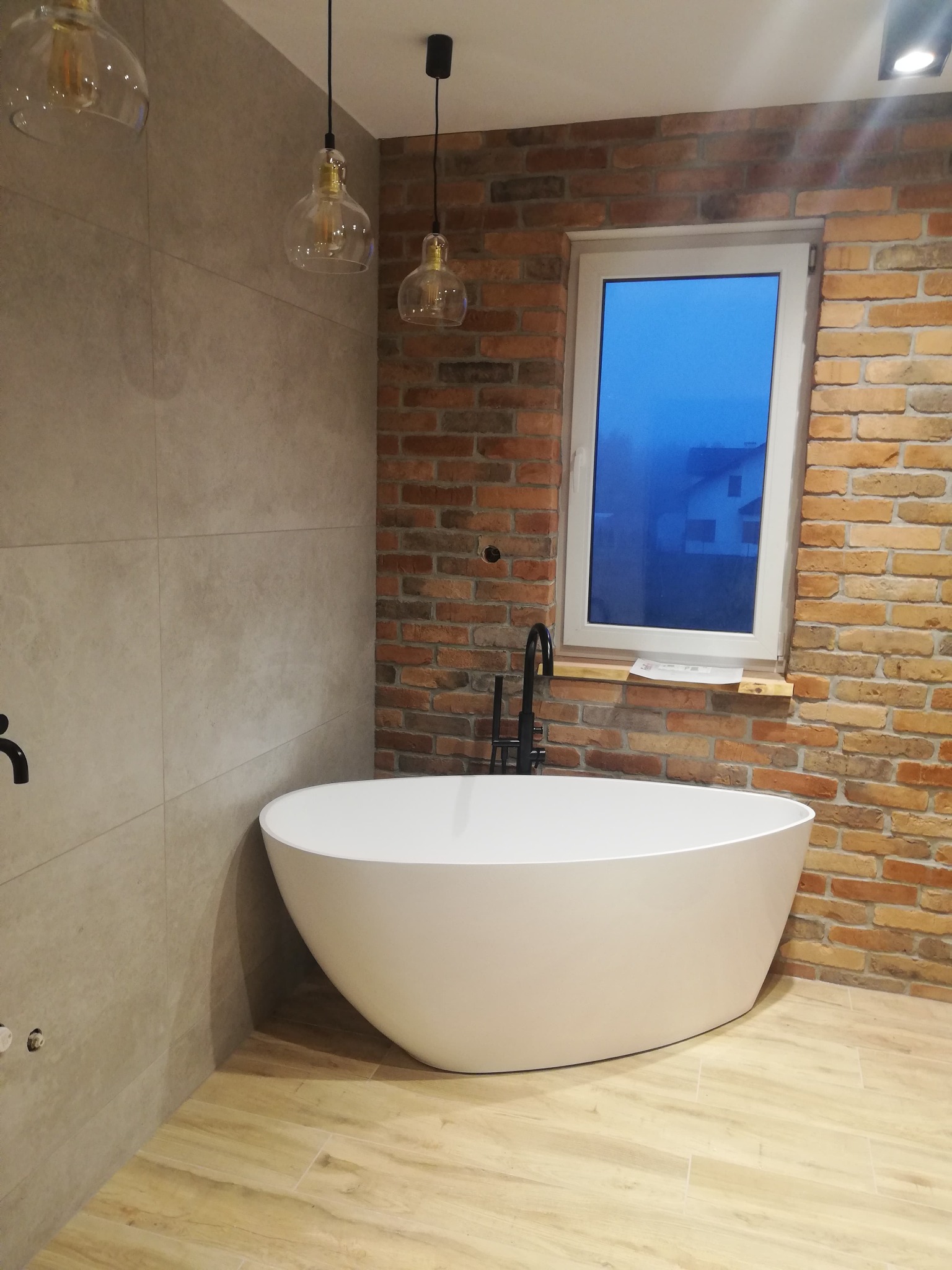 Nowoczesna łazienka z wolnostojącą białą wanną, ceglaną ścianą, okrągłymi lampami wiszącymi i widokiem na zewnątrz przez okno.