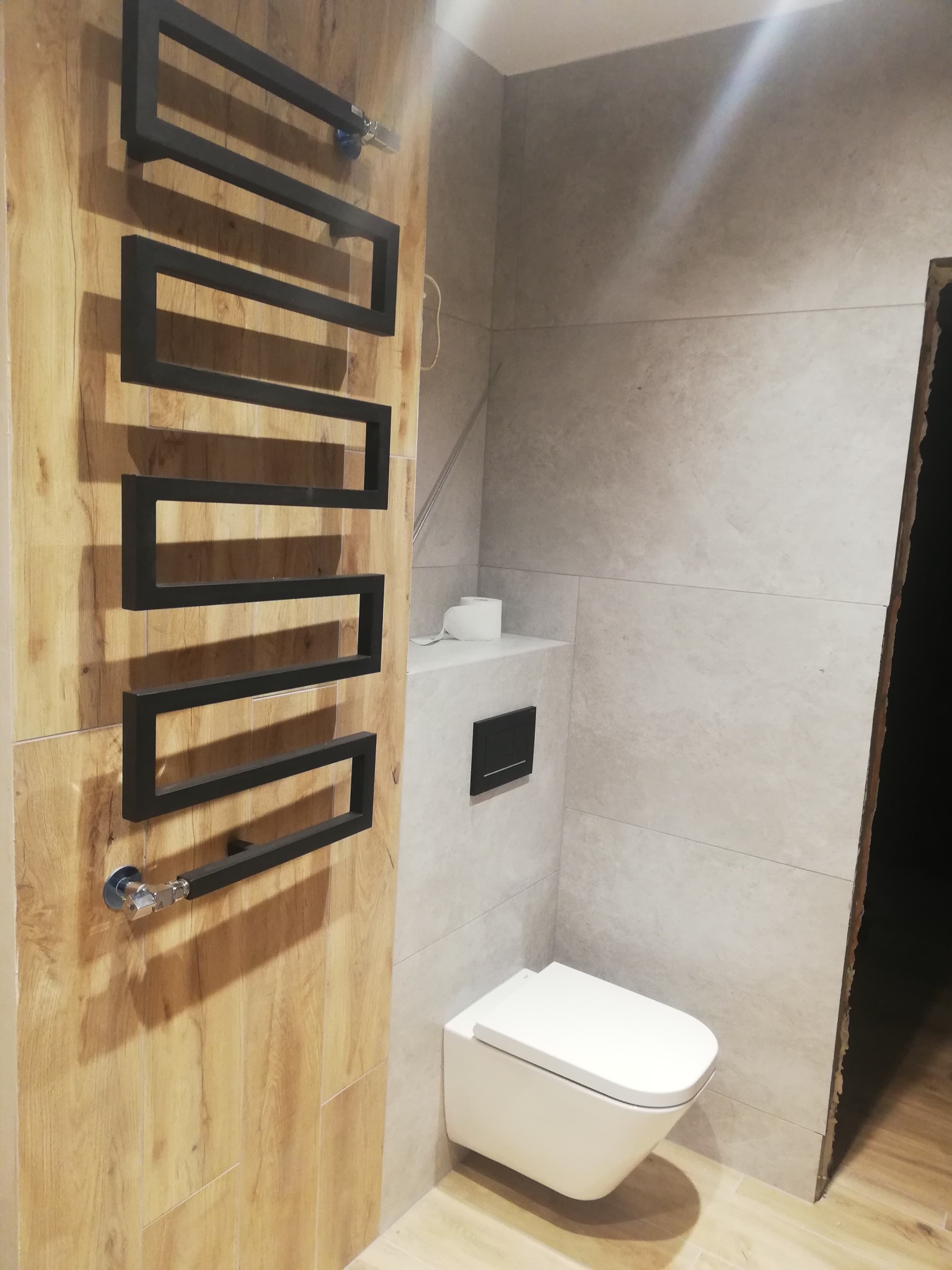 Nowoczesna łazienka z podwieszaną toaletą, grzejnikiem drabinkowym w geometrycznym stylu na drewnopodobnej ścianie i szarymi płytkami.