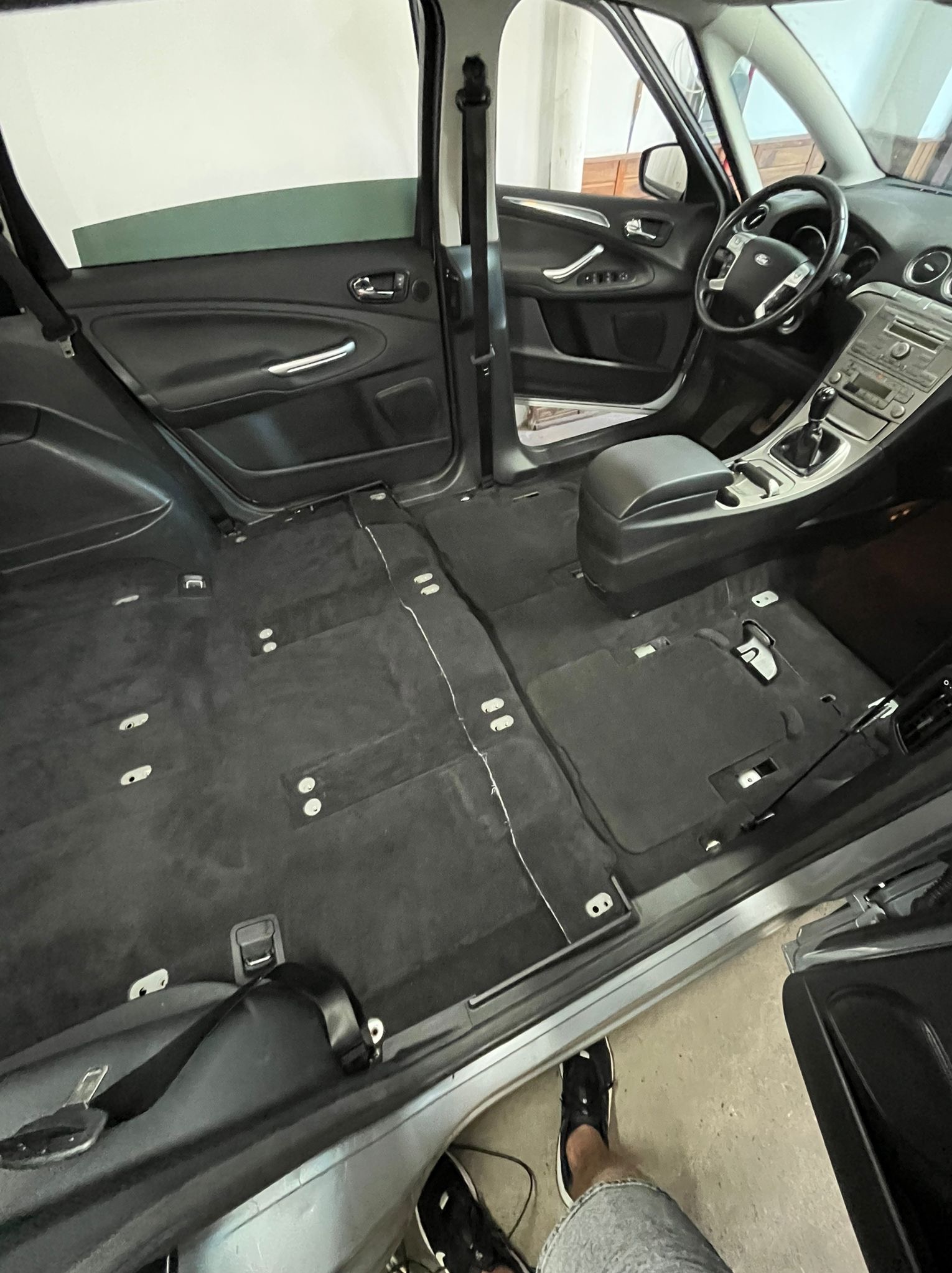 Wnętrze samochodu typu van z częściowo zdemontowaną tapicerką podłogową, widoczne elementy mocujące i pasy bezpieczeństwa, przygotowanie do czyszczenia lub naprawy podłogi.