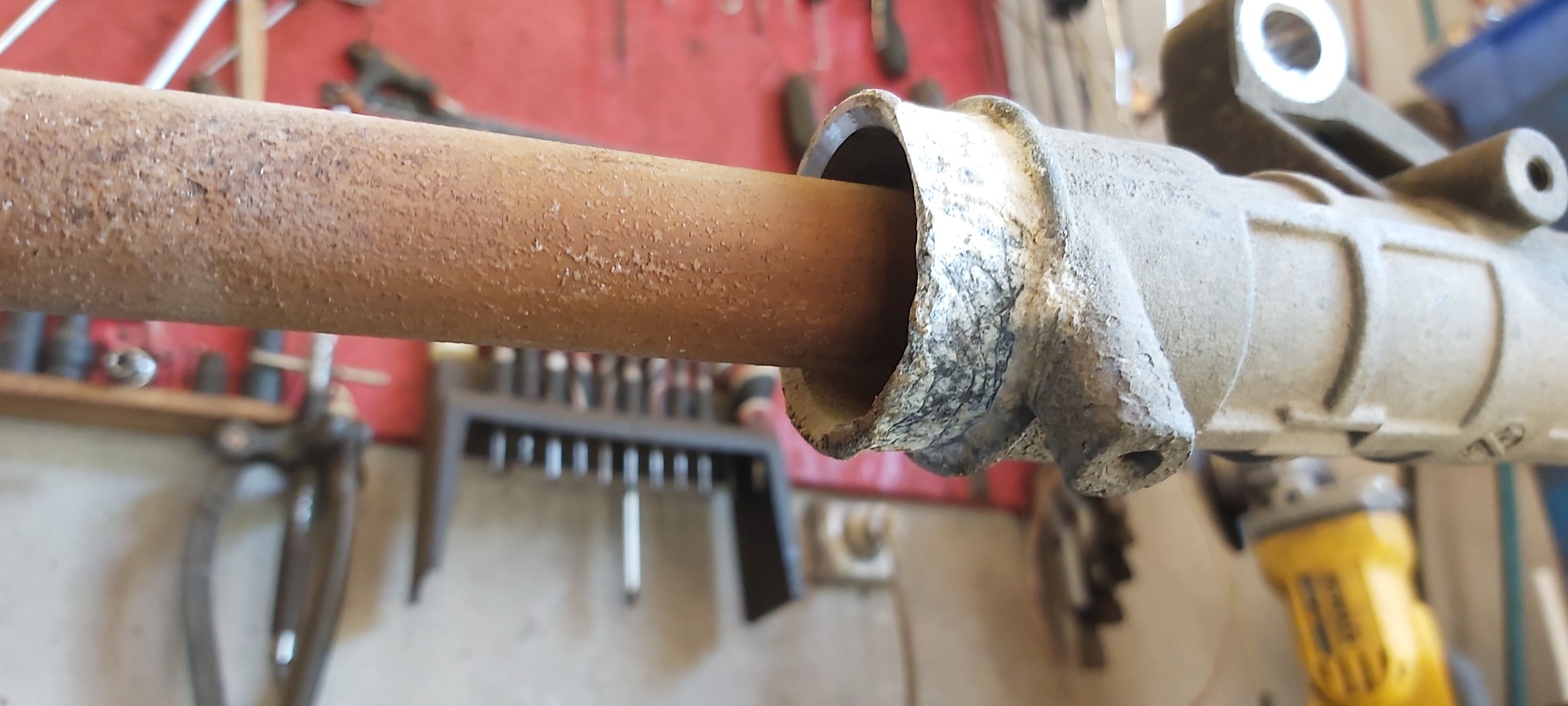 Zardzewiała rura w metalowej obudowie na tle warsztatu z narzędziami, widoczna szlifierka kątowa.