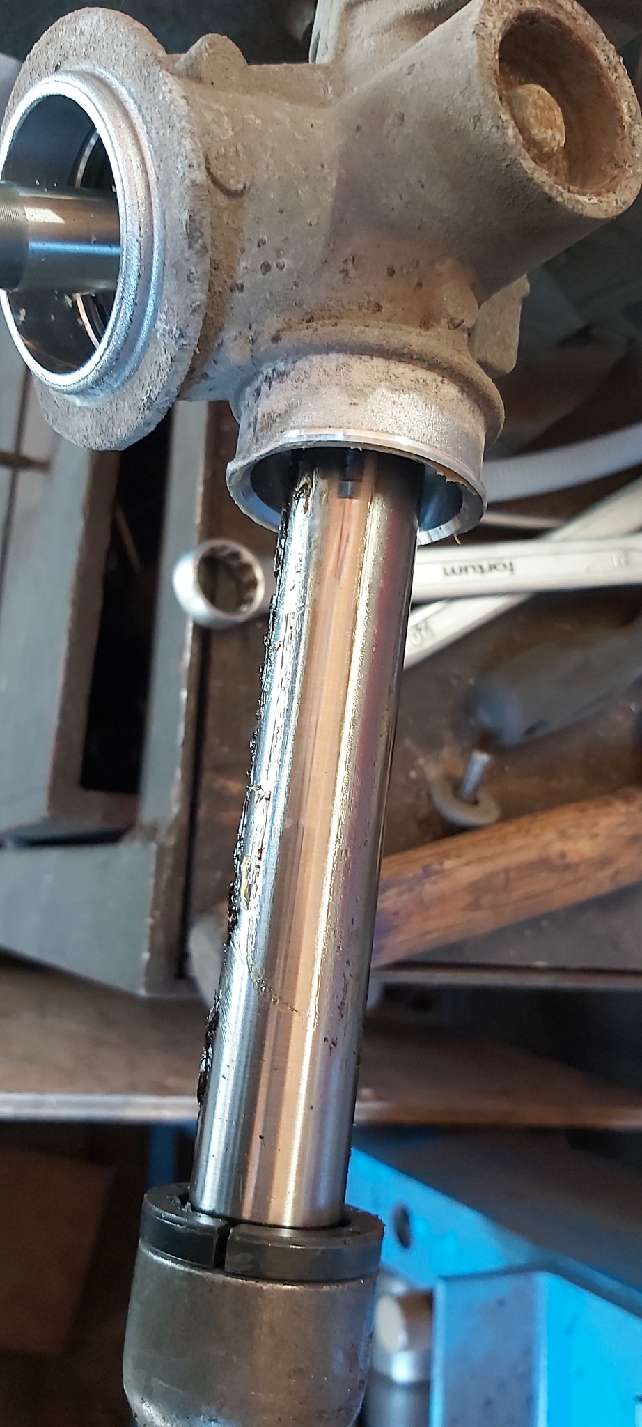 Detail of a disassembled, greasy steering rack with visible wear and tear, set against a cluttered workshop backdrop with tools.