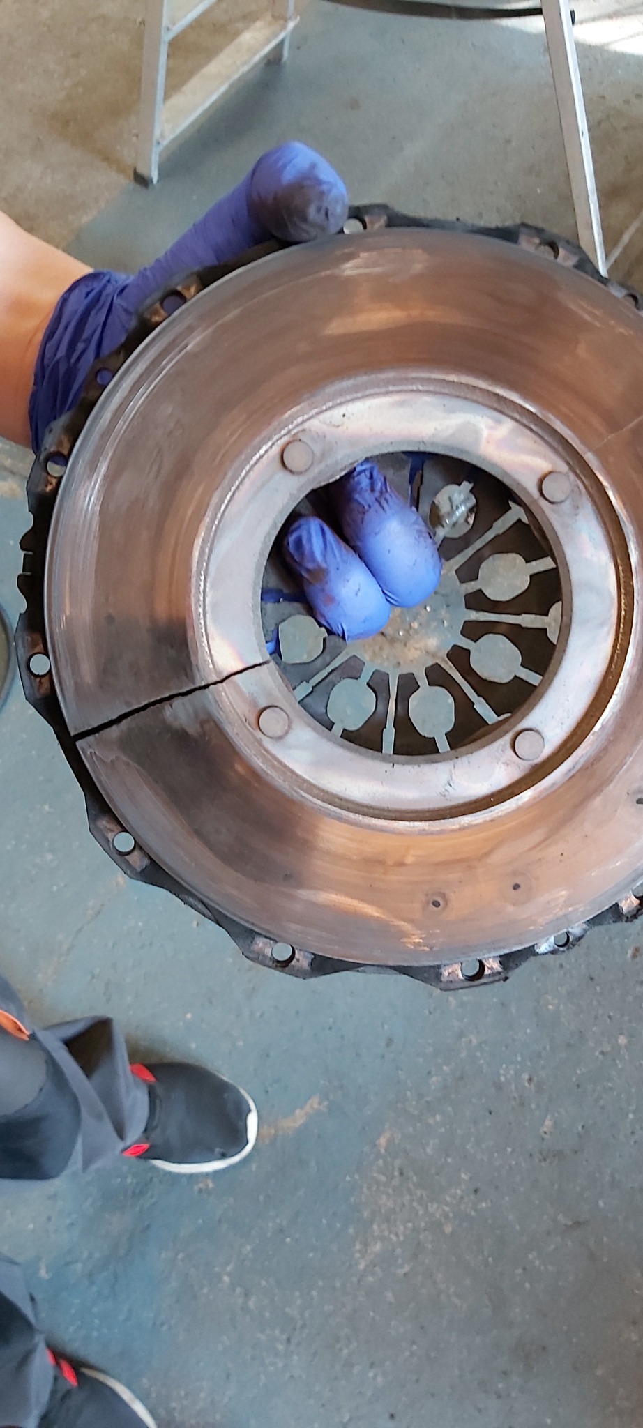 Pęknięte sprzęgło samochodowe trzymane w dłoniach w niebieskich rękawicach ochronnych, widoczne ślady zużycia i uszkodzenie mechaniczne.
