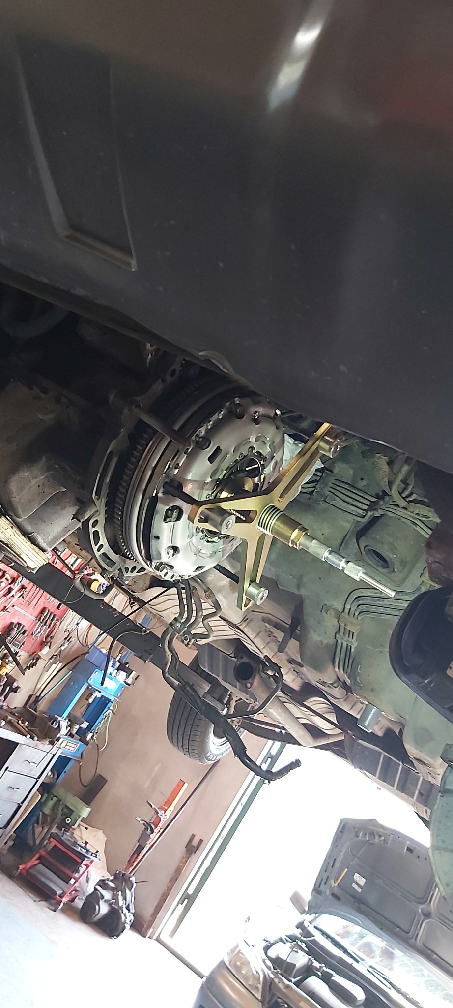 Widok od dołu na zamontowane sprzęgło samochodowe z użyciem specjalistycznego narzędzia w warsztacie, widoczne przewody i elementy podwozia.