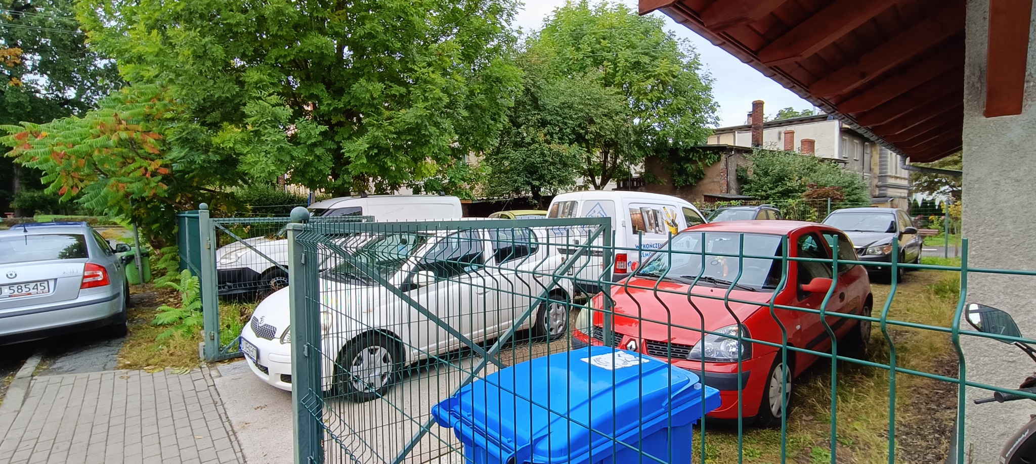 Ujęcie zza zielonego ogrodzenia na zaparkowane samochody różnych marek i kolorów, w tle budynek, drzewa i biały bus z reklamą usług wykończeniowych.