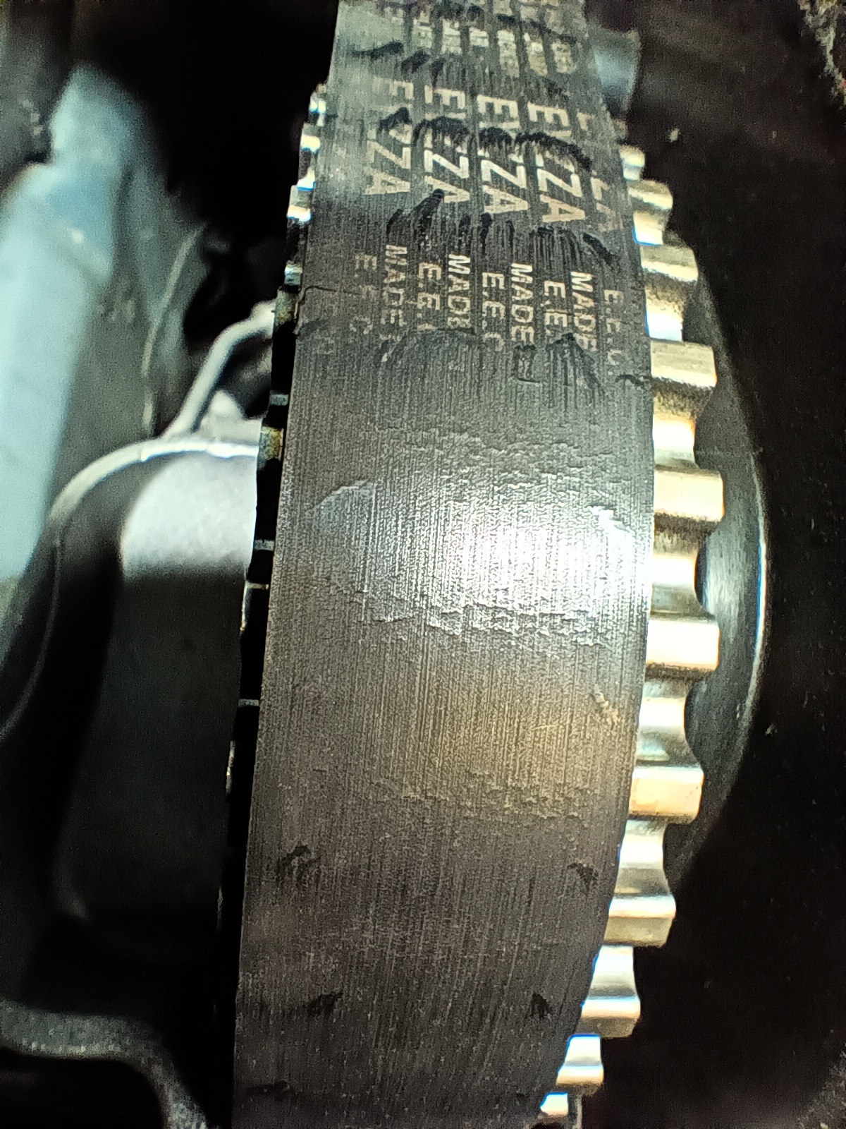 Zużyty pasek rozrządu E.E.C. E7ZA na kole zębatym w silniku samochodowym, widoczne ślady eksploatacji i otarcia.