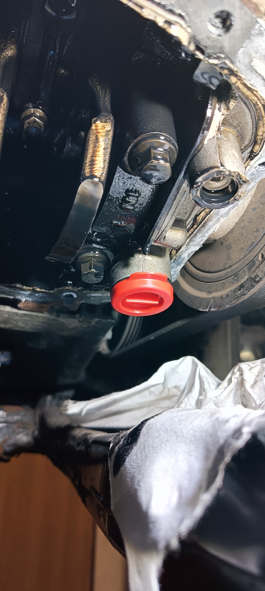 Części silnika samochodowego z czerwoną zaślepką i śladami oleju, widoczne śruby i pasek klinowy.