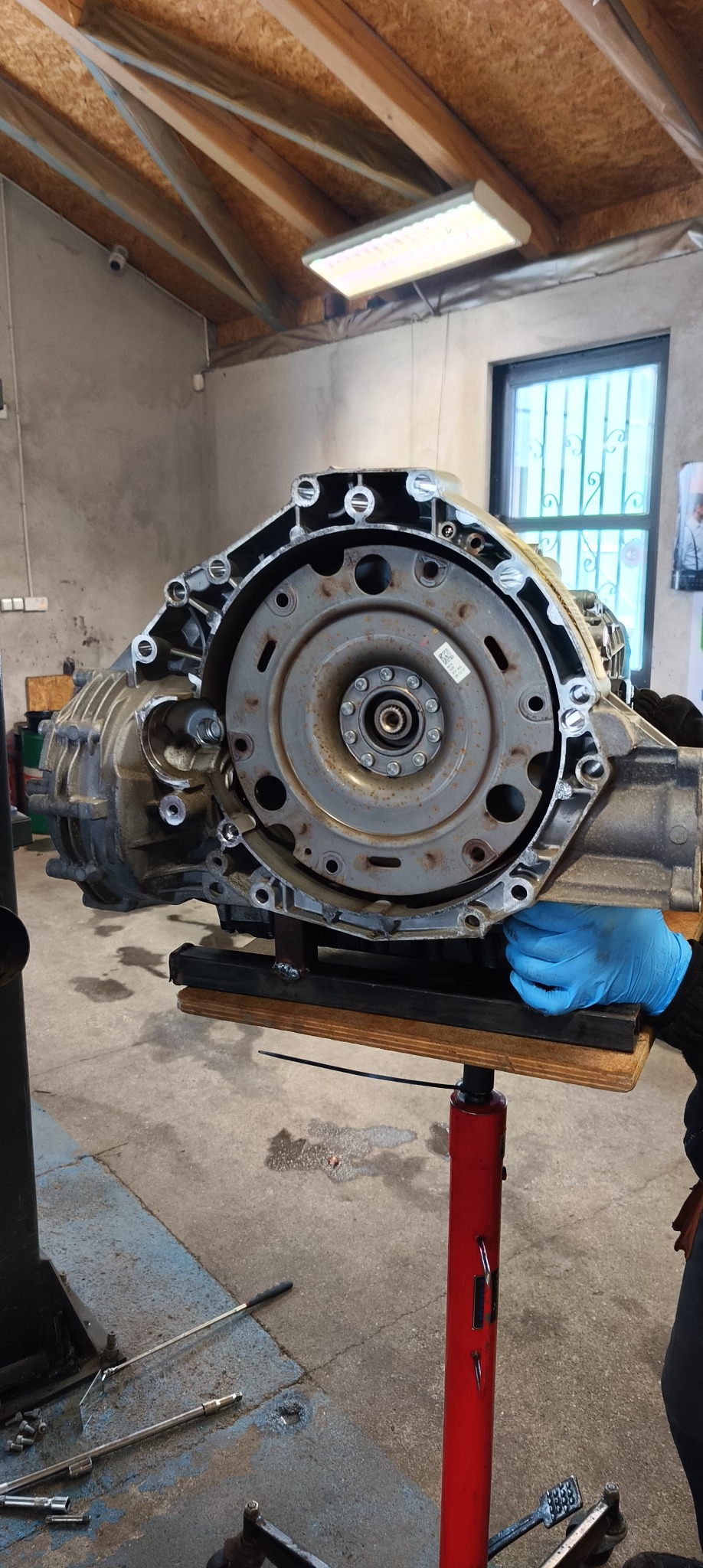 Otwarta skrzynia biegów samochodowych, podparta na czerwonym podnośniku warsztatowym, widoczna dłoń w niebieskiej rękawicy.