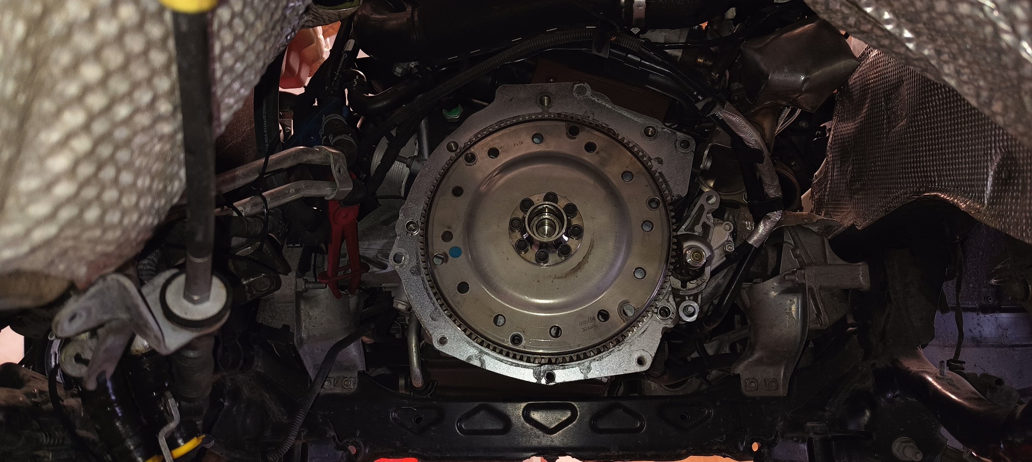 Widok od dołu na sprzęgło samochodowe podczas montażu w komorze silnika, widoczne przewody, osłony termiczne i narzędzia.