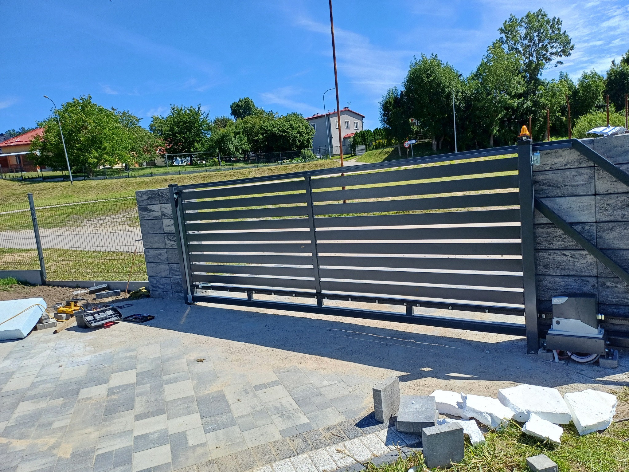 Nowoczesna, pozioma brama wjazdowa z automatyką, zamontowana na betonowych słupkach, widoczne narzędzia i materiały budowlane w trakcie montażu, kostka brukowa na podjeździe.
