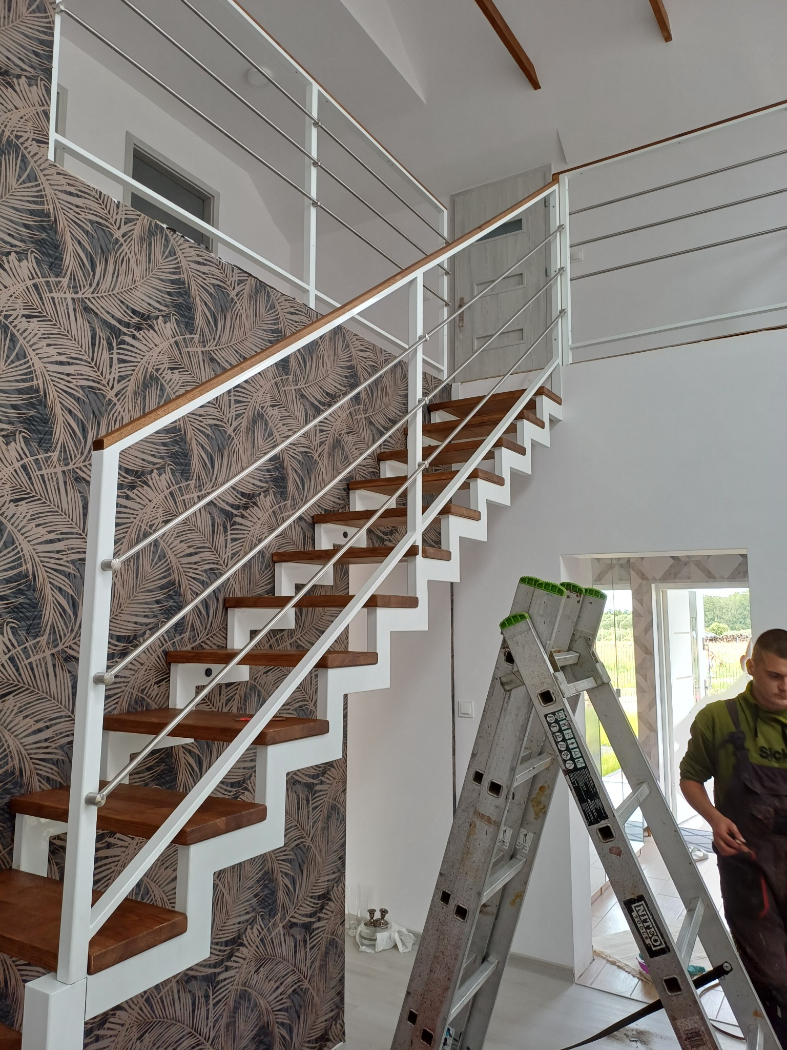 Nowoczesne schody z drewnianymi stopniami i białą konstrukcją, balustrada z nierdzewnych prętów, widoczna drabina i fragment sylwetki montera.