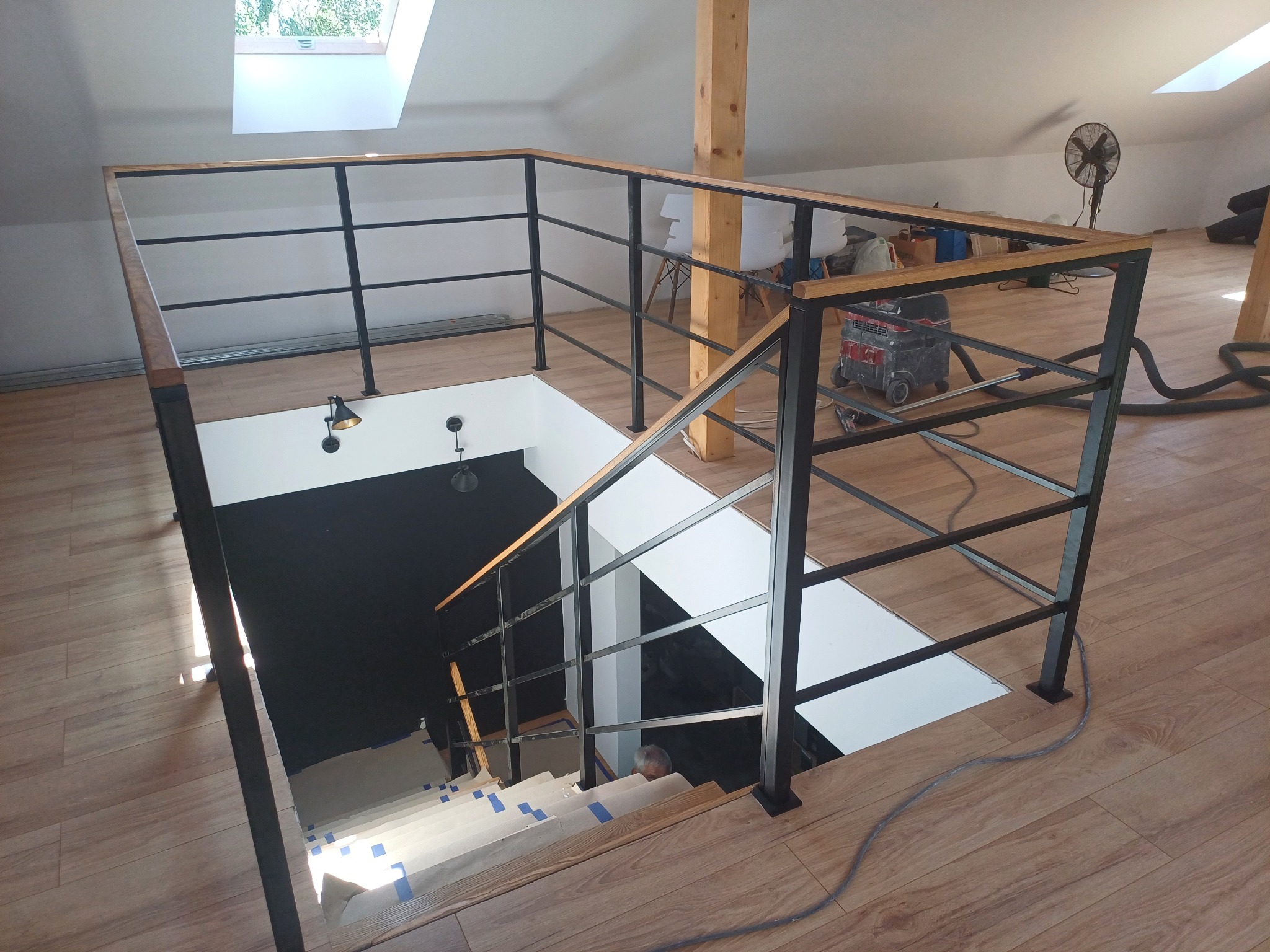 Wnętrze domu z drewnianą podłogą i metalową konstrukcją schodów z drewnianą poręczą, widoczne zabezpieczone schody w trakcie prac wykończeniowych i odkurzacz przemysłowy w tle